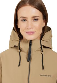 Beige wasserdichte Jacke mit hohem Kragen, schwarzem Reißverschluss und verstellbaren Kordeln. Verfügt über ein dezentes Markenlogo auf der Vorderseite.