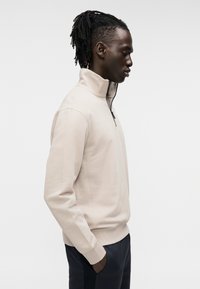 Μπεζ φούτερ με φερμουάρ στο λαιμό, μακριά μανίκια και άνετη εφαρμογή, υφασμάτινη υφή, ριμπ μανσέτες και διακριτική λεπτομέρεια ραφής. Φορεμένο με σκούρο παντελόνι.