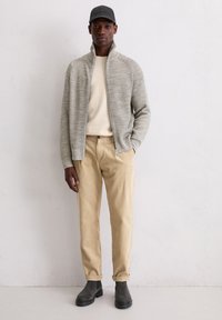 Grijze gebreide jas met hoge kraag, beige trui, beige corduroy broek en zwarte enkellaarsjes, aangevuld met een donkere pet.