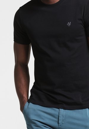 T-shirt en coton noir avec un col rond et des manches courtes, arborant un petit emblème gris sur la poitrine gauche. Associée à un short bleu clair.