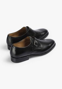 Par de zapatos formales de hombre de cuero negro con detalle de hebilla plateada y tacón bajo, exhibidos sobre un fondo blanco.