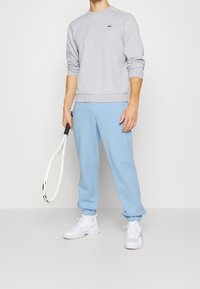 Grå sweatshirt med en liten grön logotyp, matchad med ljusblå joggers och vita sportskor, håller en tennisklubba.