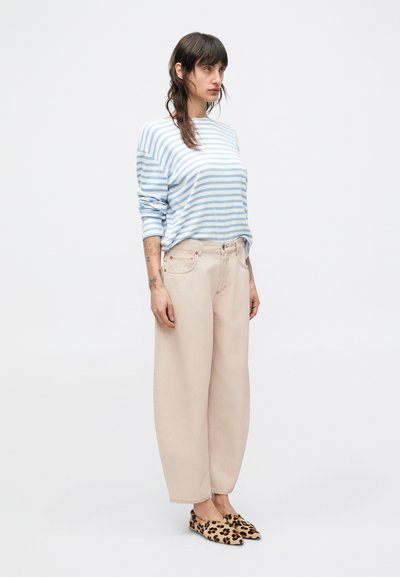 Μπλούζα με μακριά μανίκια σε ριγέ μπλε και άσπρο, μπεζ παντελόνι με φαρδιά μπατζάκια και παπούτσια slip-on με σχέδιο λεοπάρδαλης, στέκεται μπροστά σε φωτεινό φόντο.