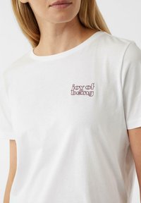 Weißes Baumwoll-T-Shirt mit kurzen Ärmeln, das auf der linken Seite einen kleinen burgunderrot gestickten Text "joy of being" trägt. Glatte Textur.