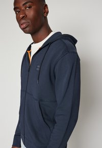 Marineblauwe zip-up hoodie met contrasterende oranje ritsdetails, katoenmix stof, voorzakken en een klein logo op de borst.