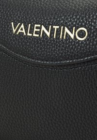 Mustat nahkakäsilaukku, jossa on kultainen "Valentino"-logo. Suunnittelussa on kaareva läppä ja hienovaraiset ommeltyylit.