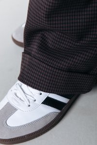 Zapatillas blancas y grises con rayas negras, fabricadas en material de ante y con cordones blancos. Se pueden ver pantalones de cuadros marrones con dobladillo.