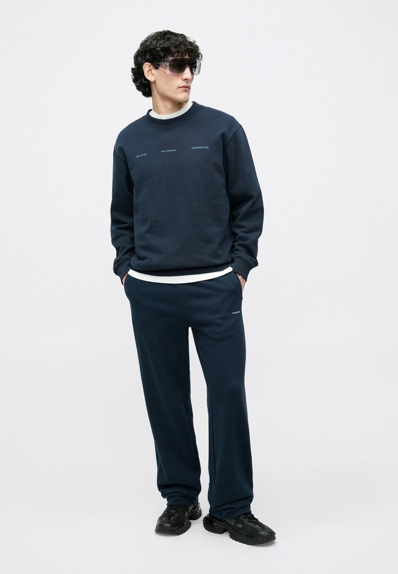 Sweat-shirt bleu marine avec des accents textuels, assorti à un pantalon de jogging bleu marine. Le mannequin porte des lunettes de soleil transparentes et des baskets noires.