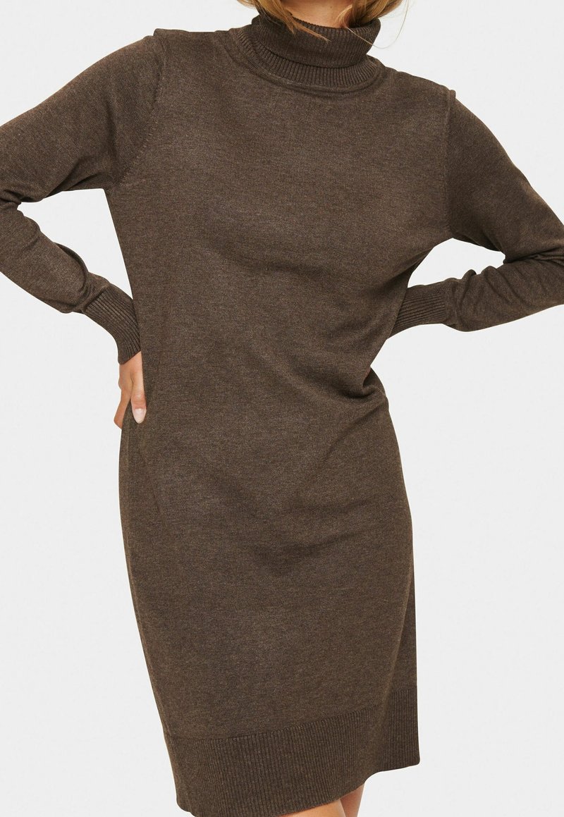 Femme portant une robe en tricot marron à col roulé et manches longues avec des poignets et un ourlet côtelés, debout les mains sur les hanches devant un fond uni.