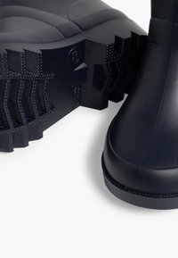 Botas de lluvia de goma negra con suela texturizada y patrones de banda de rodadura gruesos. Superficie superior lisa con punta redondeada y refuerzos en el talón.