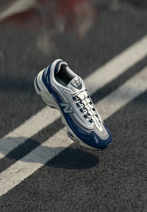 Blauer und weißer New Balance Sportschuh, der über Asphalt mit weißen Fahrbahnmarkierungen schwebt und darunter einen Schatten wirft.