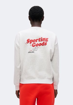 Personne portant un sweat-shirt gris clair avec texte rouge "Sporting Goods" et logo dans le dos, associé à un pantalon rouge vif, vue de dos.