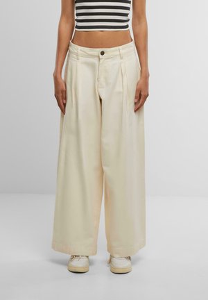 Lange broek in licht crèmekleurige stof met plooien, met een knoopsluiting en zijzakken, gecombineerd met witte sneakers.