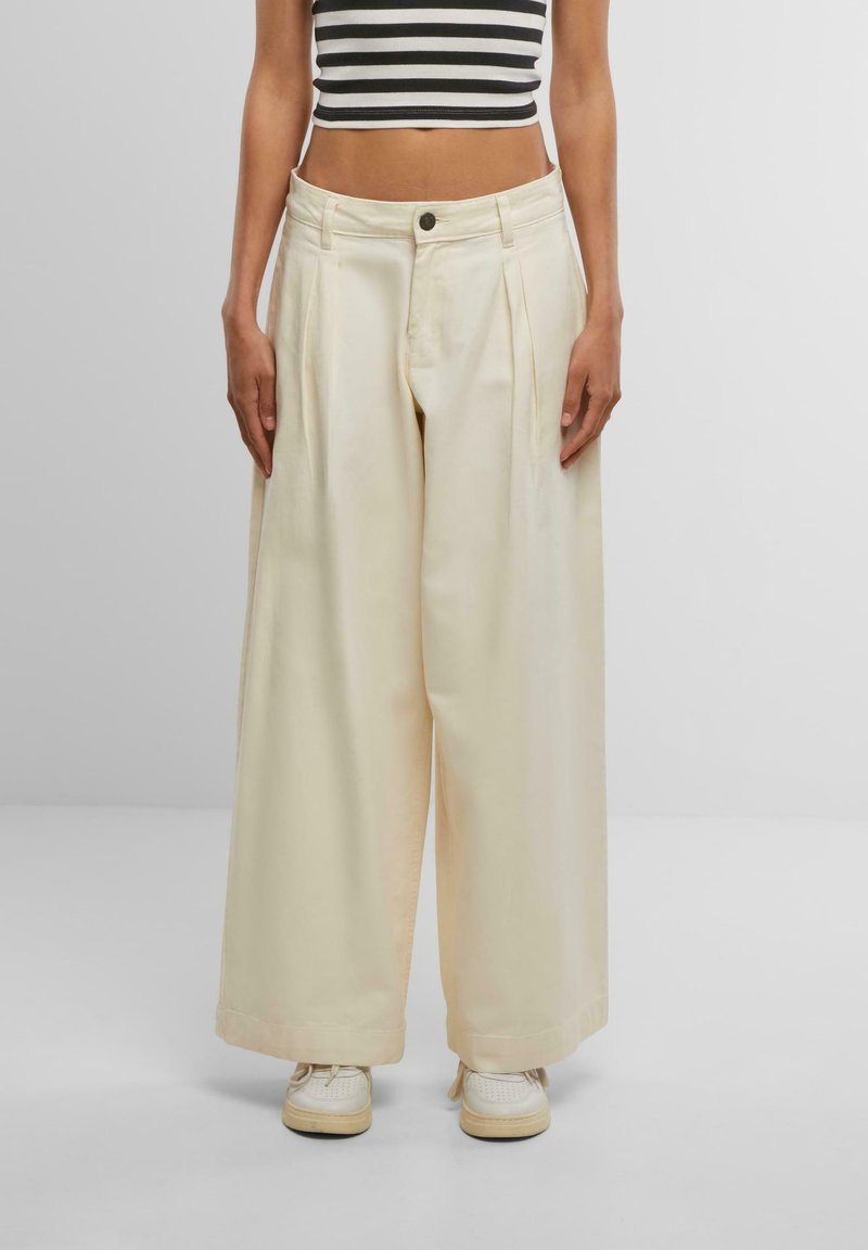 Wide-leg trousers in light cream fabric with pleats, featuring a button closure and side pockets, paired with white sneakers.
