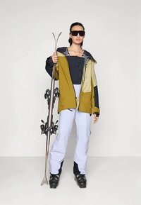 Γυναίκα με εξοπλισμό σκι κρατώντας σκι, φορώντας χρωματιστό μπουφάν, λευκά παντελόνια σκι, μαύρες μπότες σκι και μεγάλα σκούρα γυαλιά ηλίου.