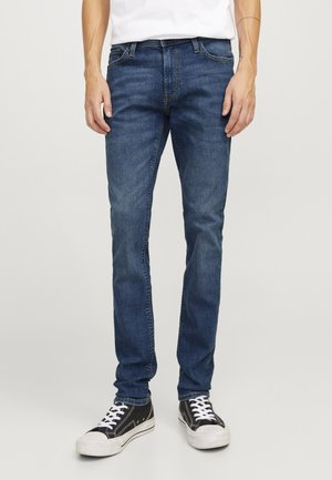 Tamne traperice u tamnoplavoj boji, uskog kroja, s klasičnim dizajnom od pet džepova i detaljima šivanja. Uparene s crnim tenisicama.