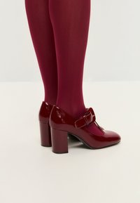Cizme roșii din piele patent cu un design în T și o cataramă decorativă. Asortate cu colanți bordeaux, având o textură netedă și strălucitoare.