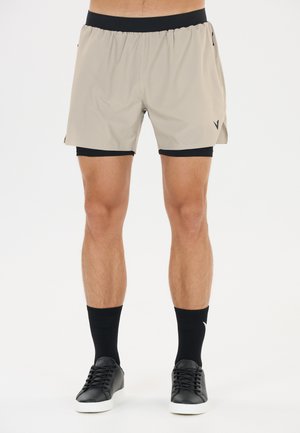 Jambes masculines portant un short de sport beige superposé, des chaussettes noires et des baskets noires avec des semelles blanches sur fond blanc.