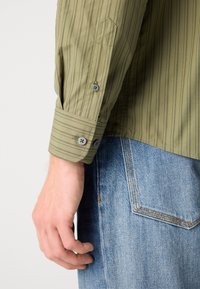 Gros plan d'une personne portant une chemise à manches longues rayée verte avec des poignets boutonnés et un jean en denim bleu avec des coutures visibles sur la poche arrière.