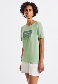 T-shirt en coton vert clair avec un col rond et des manches courtes, présentant un imprimé graphique incluant le mot "GOLDENHOUR" en texte foncé.