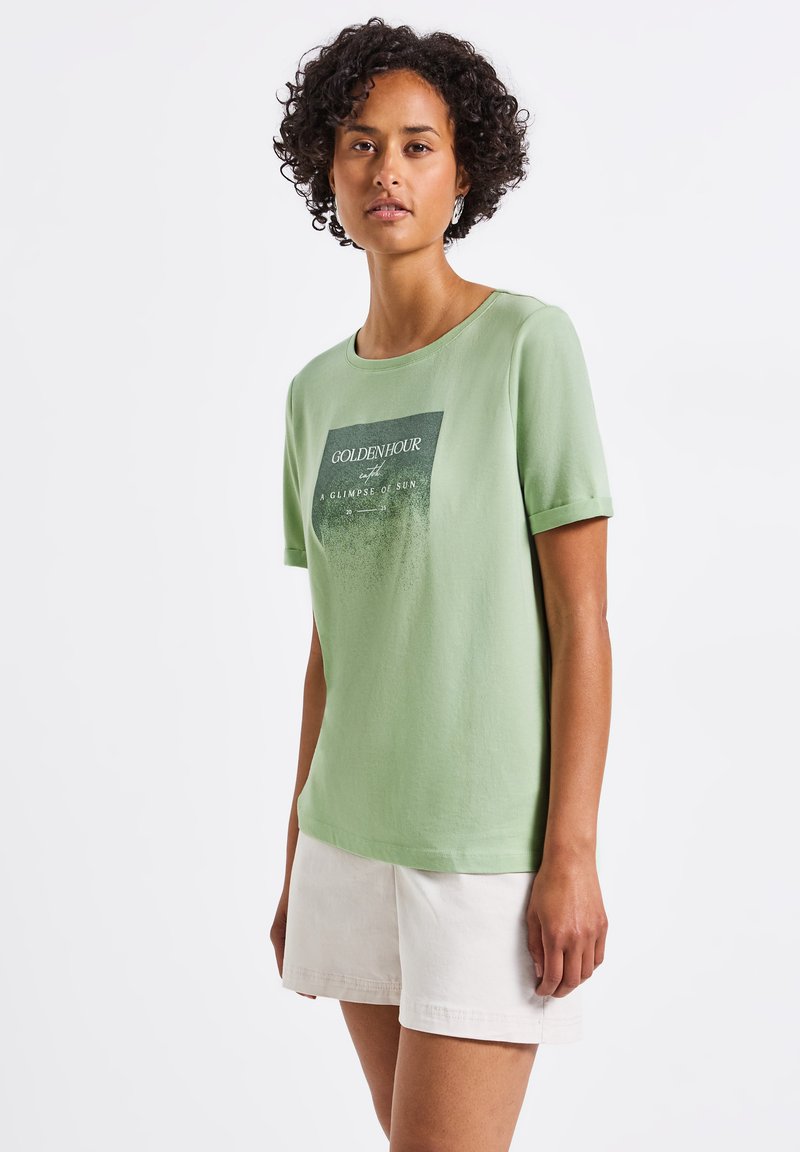 T-shirt en coton vert clair avec un col rond et des manches courtes, présentant un imprimé graphique incluant le mot "GOLDENHOUR" en texte foncé.