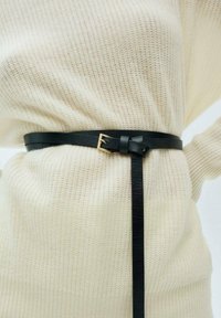 Cintura in pelle nera con fibbia in metallo dorato, caratterizzata da un dettaglio annodato e una lunga coda, indossata sopra un maglione di lana beige chiaro.