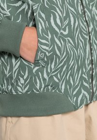 Green zip-up jacket with a leaf pattern in light green. Ribbed cuffs and hem, featuring a side pocket and a small brand tag.