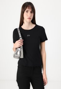 T-shirt noir à manches courtes avec le logo GAP, associé à un sac bandoulière métallisé argent. Le tissu semble lisse et la coupe est décontractée.