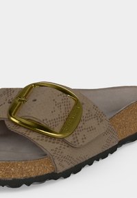 Κοντινή λήψη μιας μπεζ σαγιονάρας Birkenstock με υφή στο λουράκι, μεγάλο χρυσό αγκράφα, φελλό πάτο και μαύρη προοδευμένη σόλα.