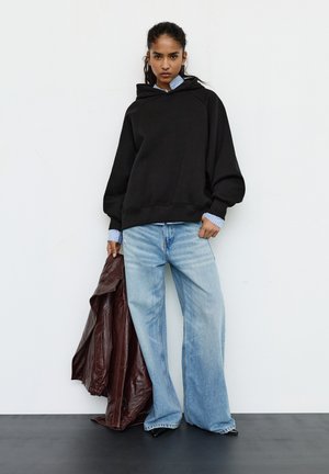 Zwarte oversized hoodie met capuchon, wijde mouwen en geribbelde boorden, gecombineerd met lichtblauwe uitlopende jeans. Model houdt een bruine leren jas vast.