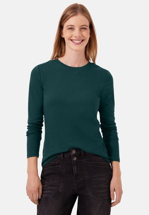 Lächelnde Frau mit glattem Haar trägt ein dunkelgrünes Langarmshirt und hoch taillierte schwarze Jeans vor einfarbigem Hintergrund.