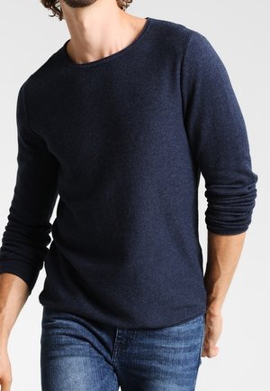 Pull à manches longues bleu marine avec un col rond et un tissu texturé. Porté avec un jean bleu clair, affichant une coupe décontractée.