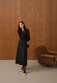 Femme en longue robe noire, gants noirs et talons beiges, debout près d'une chaise en cuir marron contre un mur en bois.