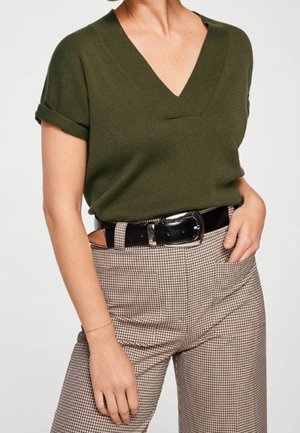 Mujer con camiseta de manga corta color verde oliva y escote en V, metida dentro de pantalones de cintura alta a cuadros con cinturón negro, mano apoyada en el bolsillo.