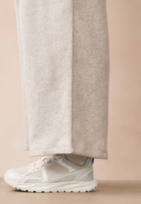 Gaiši pelēkas sporta bikses no mīksta auduma kontrastē ar krēmkrāsas sporta apaviem, kuriem ir fakturēts tīkls augšdaļā un gaiši zili akcenti.