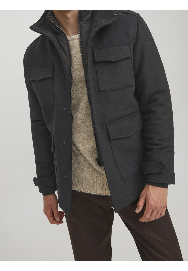 Černý kabát ze směsi vlny se zipem uvnitř, čtyřmi předními kapsami a vysokým límcem. Nosený přes béžový pletený svetr a tmavé kalhoty.