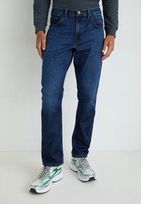 Jeans de mezclilla azul oscuro con un corte recto, que cuentan con bolsillos frontales, cremallera y cierre de botón metálico. Llevados con zapatillas grises y verdes.