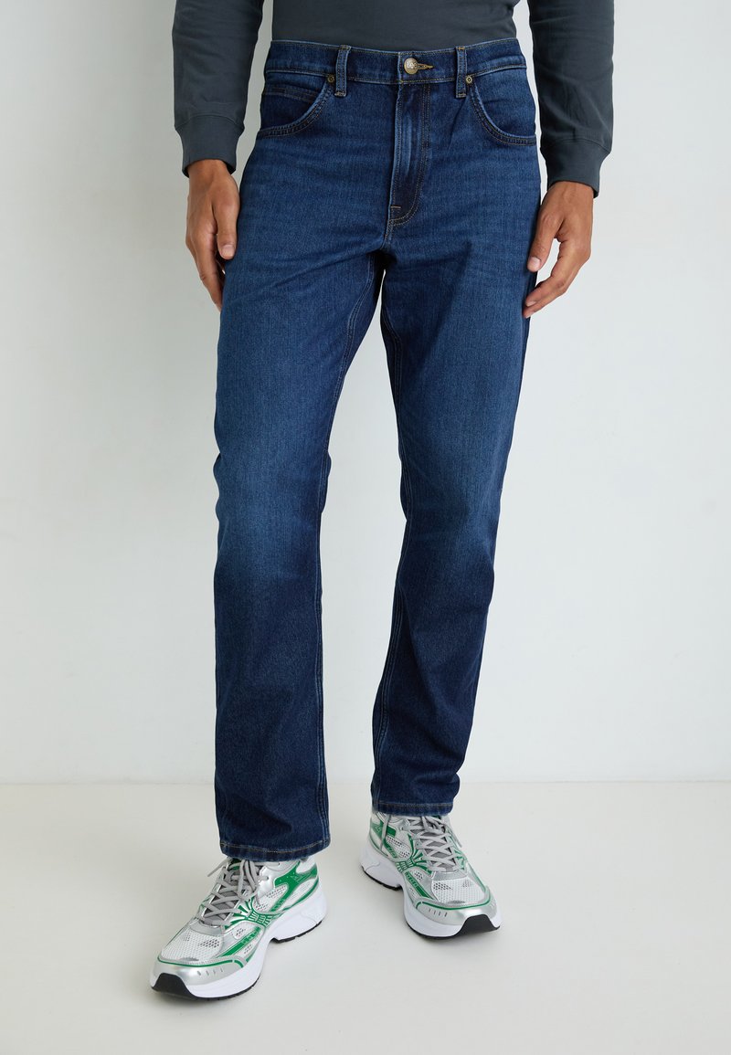 Jeans de mezclilla azul oscuro con un corte recto, que cuentan con bolsillos frontales, cremallera y cierre de botón metálico. Llevados con zapatillas grises y verdes.