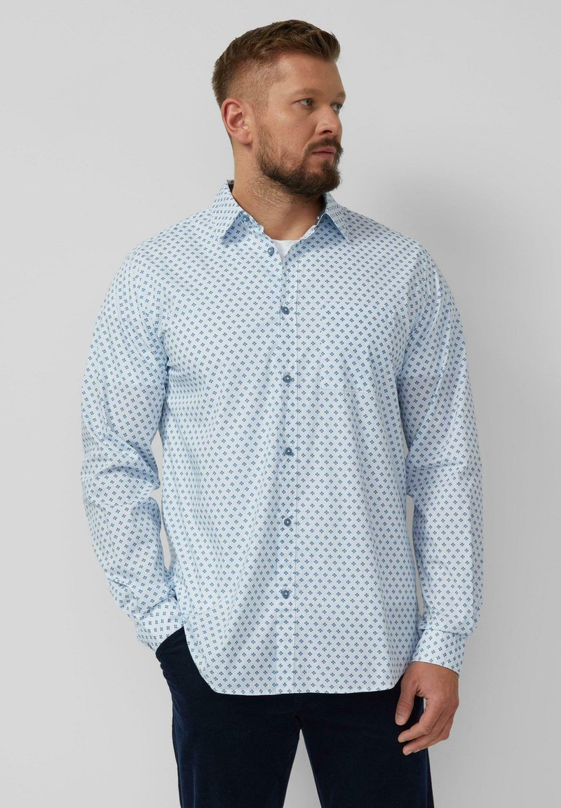 Man met kort haar en baard, gekleed in een lichtblauw overhemd met patroon en donkere broek, staand met één hand in zijn zak.