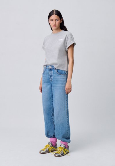 T-shirt grigio a maniche corte, jeans a gamba larga a vita alta azzurri chiari, scarpe da ginnastica argento con lacci gialli e calzini rosa, in piedi davanti a uno sfondo neutro.