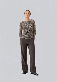 Frau steht mit den Händen in den Taschen, trägt ein langärmliges Oberteil mit Leopardenmuster, dunkelbraune weit geschnittene Hose und schwarze Schuhe.