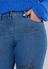Denim jeans in medium blue met een slim fit. Heeft zakken en bedrukte tekstontwerpen in het zwart aan de voorkant en zijkant.
