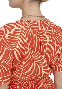 Robe rouge et beige avec un motif de feuilles, présentant des manches courtes, une taille froncée et une fermeture à bouton dans le dos.