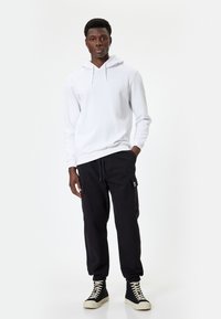 Hvid hættetrøje med snoretræk, sorte cargo bukser med sidelommer, og sorte høj-top sneakers med hvide såler. Enkel, afslappet design.