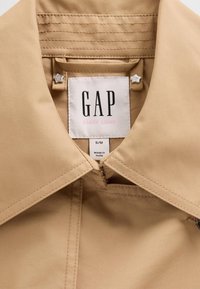 Beige jacka med krage och slät textur, med en GAP-tag, rosa detaljer och stjärnformade metallbeslag nära kragen.