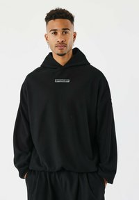 Sudadera negra de gran tamaño hecha de tejido texturizado, con un parche de logo en el frente y capucha con cordones. Se lleva con pantalones negros a juego.