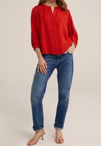 Red woven blouse with a round neckline and front buttons, paired with light blue relaxed-fit jeans and beige heeled sandals.