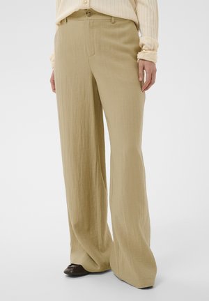 Osoba nosiaca voľné, béžové nohavice s širokými nohami v kombinácii s tmavými topánkami a svetlým, rebrovaným tričkom s dlhým rukávom, stojaca na jednoduchom pozadí.