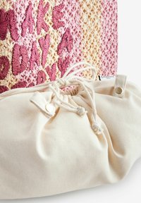 Bolsa tejida con texto bordado en rosa y crema. El cierre con cordón de algodón en la parte inferior cuenta con un detalle de botón para un ajuste seguro.