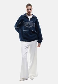 Granatowy, oversize'owy sweatshirt zapinany na zamek z białym napisem "NEWYORK" oraz stójką, zestawiony z białymi spodniami w kształcie dzwonów i białymi butami.